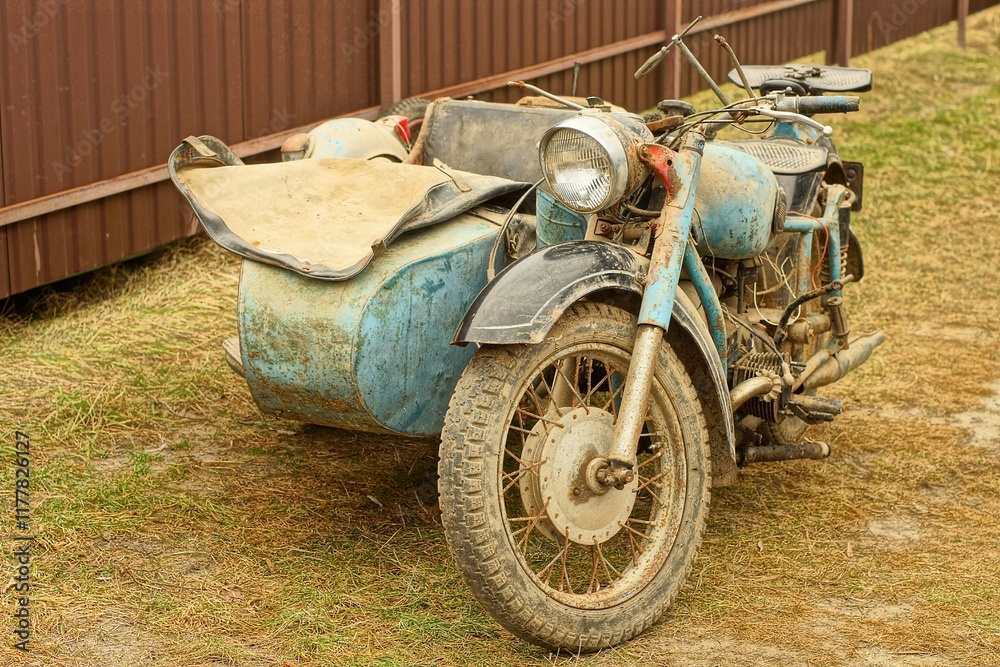 Fototapeta premium one retro broken blue military iron old with engine and two wheels industrial dirty motorcycle with a sidecar stands on the ground on the street near the brown fence during the day