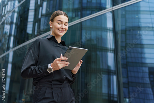 Young woman in black attire using tablet outdoors in urban setting with modern glass buildings