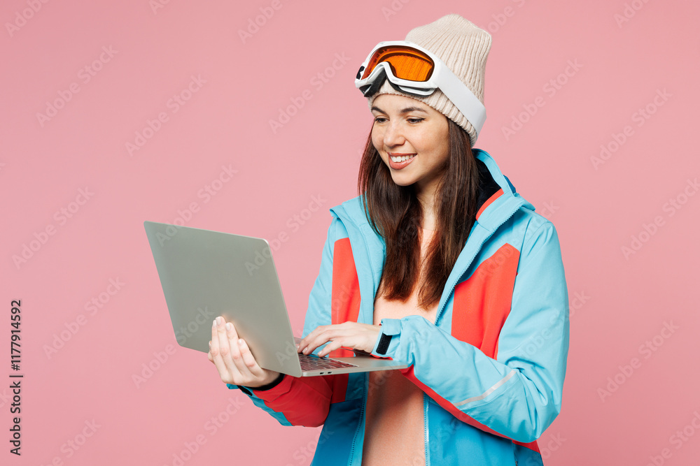 Skier young IT woman wear warm blue padded jacket ski goggles mask spend extreme weekend winter season in mountains hold use work on laptop pc computer isolated on plain pink background Hobby concept Skier young IT woman wear warm blue padded jacket ski goggles mask spend extreme weekend winter season in mountains hold use work on laptop pc computer isolated on plain pink background Hobby concept