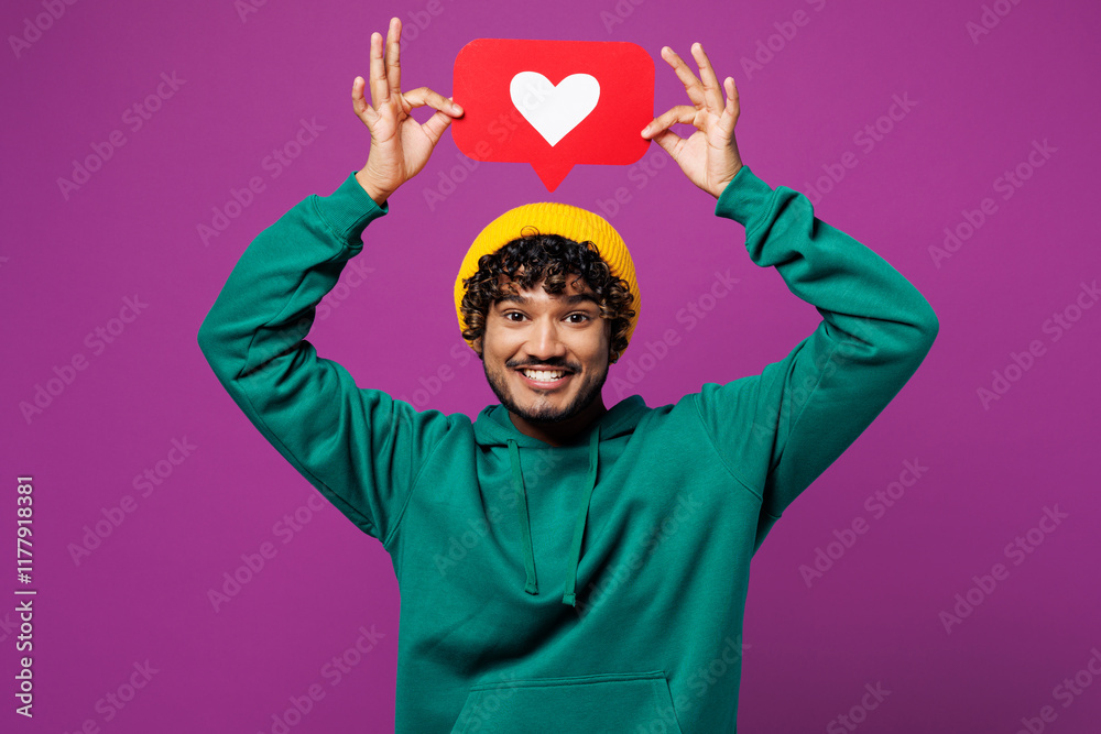 © ViDi Studio - Young happy Indian man wear green hoody yellow hat casual clothes hold heart form like icon sign from social network media feedback above head isolated on plain purple background. Lifestyle concept.