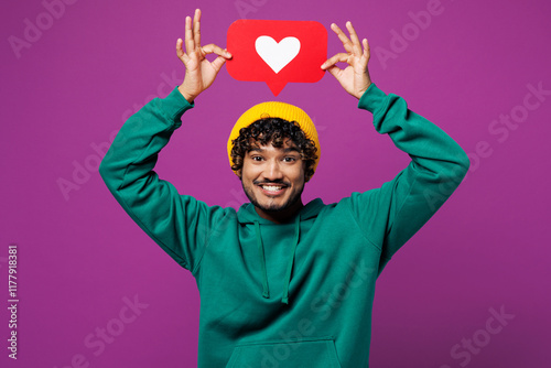 Young happy Indian man wear green hoody yellow hat casual clothes hold heart form like icon sign from social network media feedback above head isolated on plain purple background. Lifestyle concept. photo