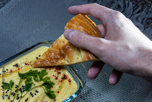 A close-up of a man's hand with a flatbread, the edge of the flatbread is immersed in a glass bowl with homemade hummus sprinkled with sesame seeds, parsley and paprika. photo