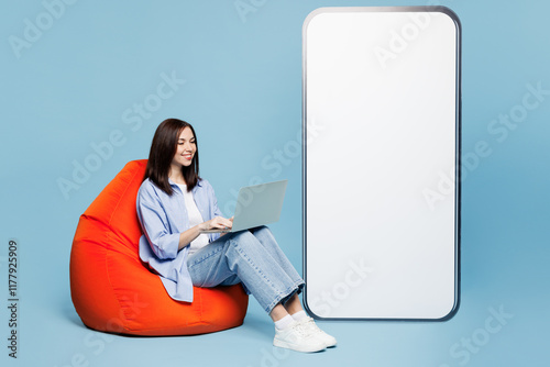 Full body young IT woman wears shirt casual clothes sit in bag chair big huge blank screen mobile cell phone smartphone with area hold use work on laptop pc computer isolated on plain blue background. photo