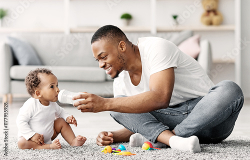 Caring young black dad giving bottle with water to his little baby at home, loving african american father babysitting cute adorable toddler child, playing together on floor in living room