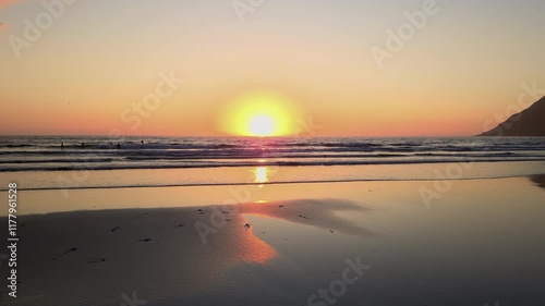 Warm sunlight illuminates the tranquil scene of a wet sandy beach in Lofoten, Norway, as the setting sun casts a golden reflection on the water's surface