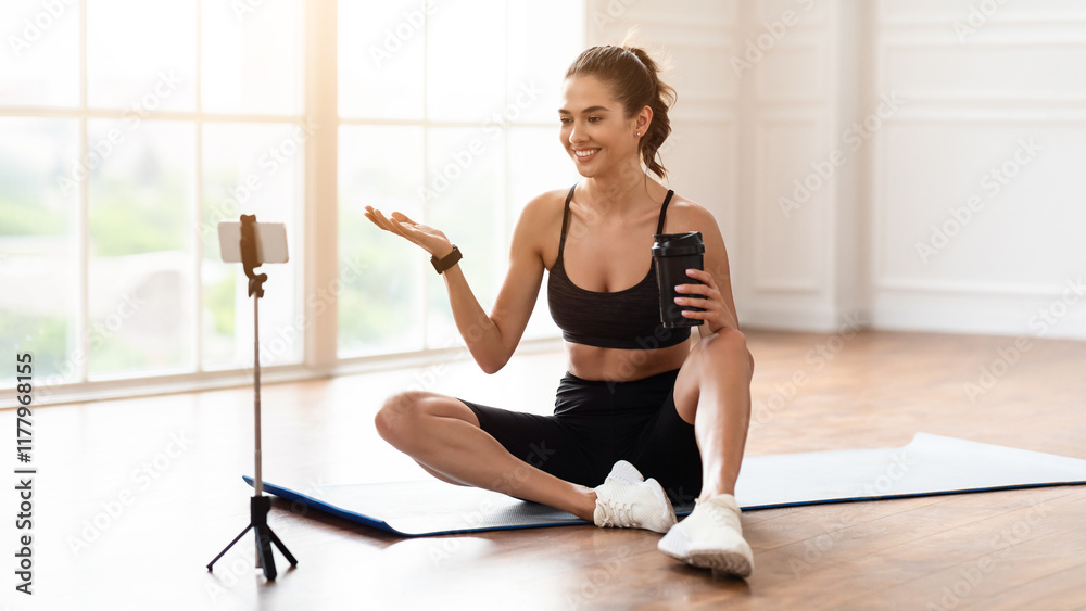 Fototapeta premium Sports Blogger, Online Coach and Remote Training, Yoga Practice. Happy millennial female sitting on mat and talking with followers, looking at webcam on mobile phone on tripod, holding shaker