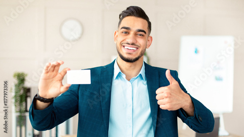 Fotografie Happy Arab guy entrepreneur holding empty business card, showing thumb up gesture at company office, mockup for design