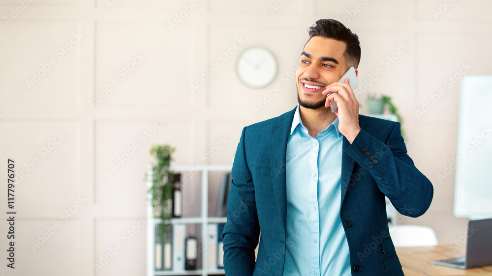 Positive young Arab man in office suit talking on mobile phone at workplace. Handsome millennial CEO having business conversation with partner or co-worker on cellphone Positive young Arab man in office suit talking on mobile phone at workplace. Handsome millennial CEO having business conversation with partner or co-worker on cellphone