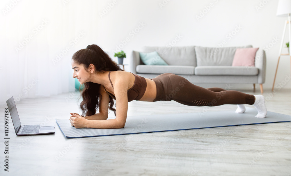 Sporty Indian woman standing in elbow plank, following video tutorial on laptop at home. Side view of Asian lady exercising her abs core muscles. Online trainings during covid lockdown Sporty Indian woman standing in elbow plank, following video tutorial on laptop at home. Side view of Asian lady exercising her abs core muscles. Online trainings during covid lockdown