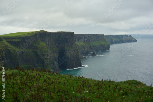 A dramatic view of the Cliffs of Moher in Ireland, with towering rocky cliffs covered in lush greenery overlooking the Atlantic Ocean. The overcast sky and misty atmosphere enhance the rugged land