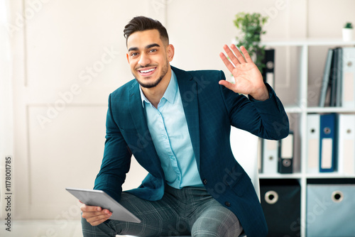 Portrait of cheerful Arab office worker in buiness suit holding tablet computer and waving at camera in modern office. Millennial guy manager using modern gadget for work, using touch pad photo