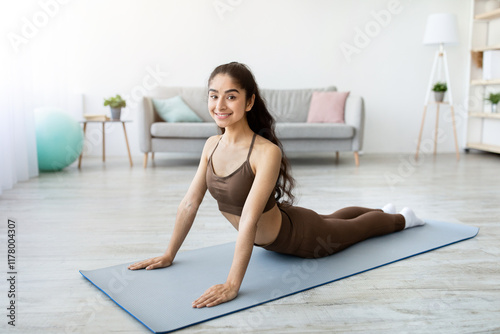 Fit young Indian woman practicing yoga, making cobra pose at home, full length. Cheerful Eastern lady having domestic training, doing flexibility exercises, enjoying active lifestyle