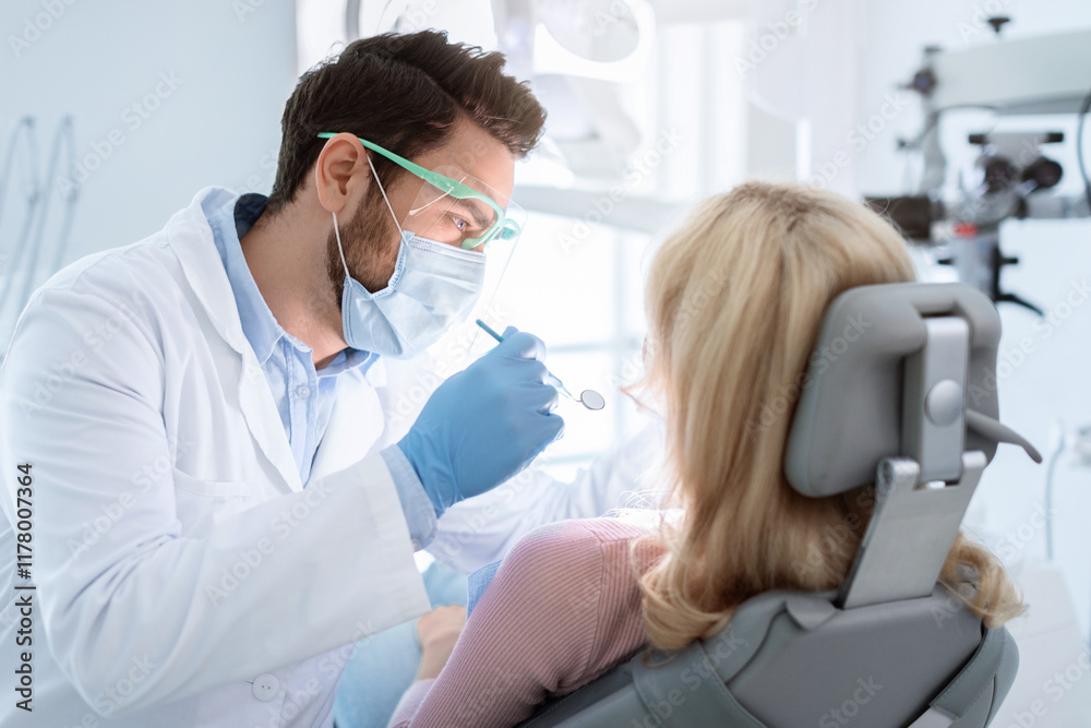 Man dentist in face mask and glasses doing treatment for patient blonde lady, holding dental tools, wearing rubber gloves. Stomatology, dentistry, modern dental clinic concept Man dentist in face mask and glasses doing treatment for patient blonde lady, holding dental tools, wearing rubber gloves. Stomatology, dentistry, modern dental clinic concept