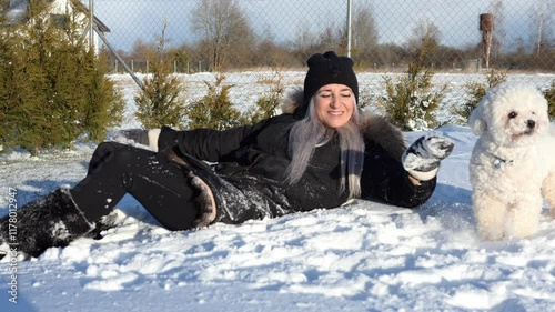Smiling young woman in black beanie and winter clothes lies in fresh snow, playing with adorable white bichon frise dog. Sunny winter day highlights joy and connection in snowy backyard moments 