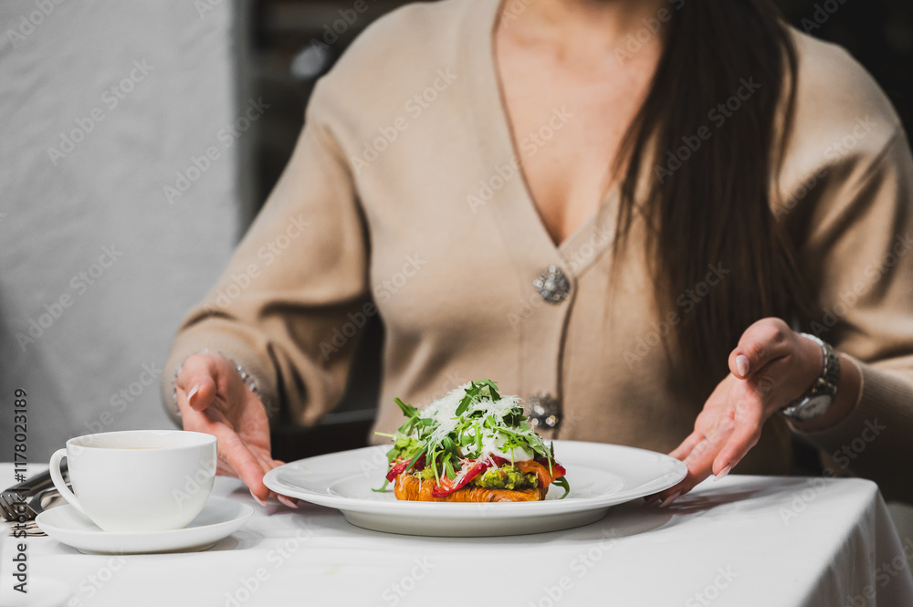 A woman presents a beautifully plated dish with greens on a white tablecloth. A cup of coffee sits beside her, adding to the cozy dining atmosphere. A woman presents a beautifully plated dish with greens on a white tablecloth. A cup of coffee sits beside her, adding to the cozy dining atmosphere.