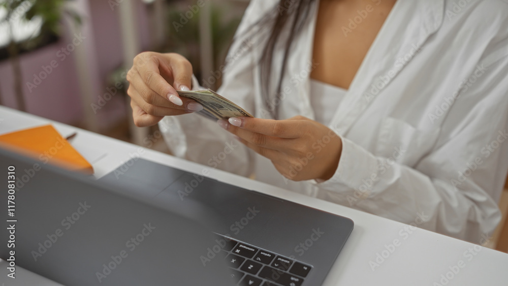 Woman counting money in a home decor environment while using a laptop and sitting at a table, showcasing both finance and lifestyle elements.