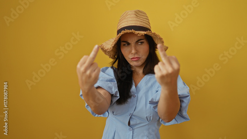 Young woman in a blue dress and straw hat making offensive gesture isolated over yellow background conveying frustration or anger photo