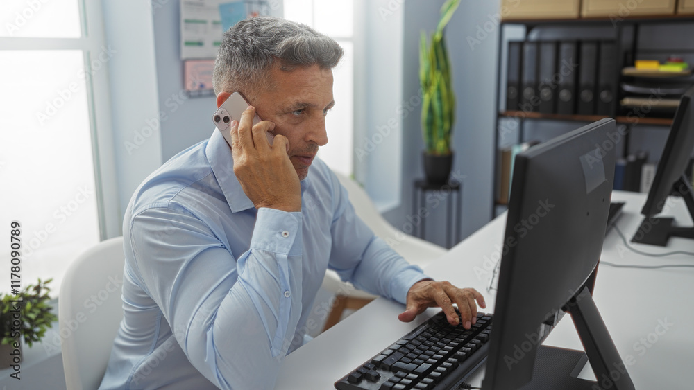 Middle-aged man talking on a phone while working on a computer in an office setting. Middle-aged man talking on a phone while working on a computer in an office setting.
