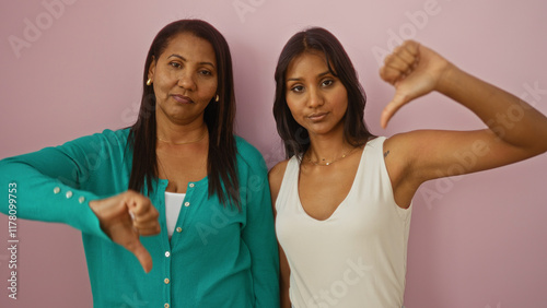 Mother and daughter showing disapproval with thumbs down over a pink background, women together displaying negative emotion