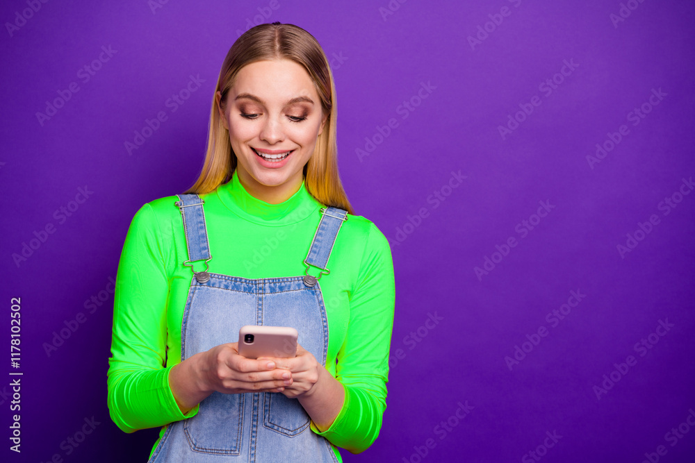 Portrait of cheerful youth holding device wearing yellow turtleneck denim jeans overalls isolated over violet purple background Portrait of cheerful youth holding device wearing yellow turtleneck denim jeans overalls isolated over violet purple background