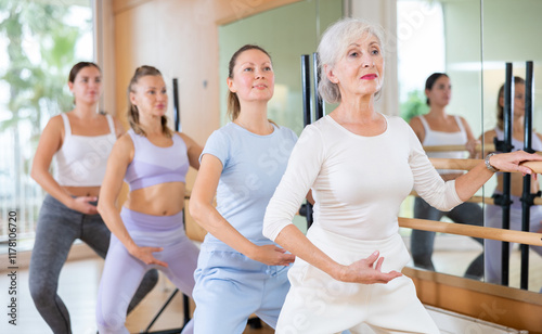 Slim aged woman and other attendees practicing ballet at ballet barre in dance studio during training session