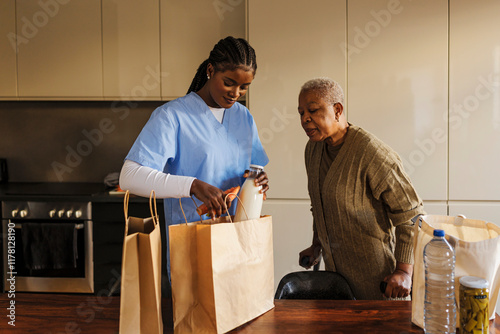 Caregiver unpacking groceries for elderly woman in kitchen photo