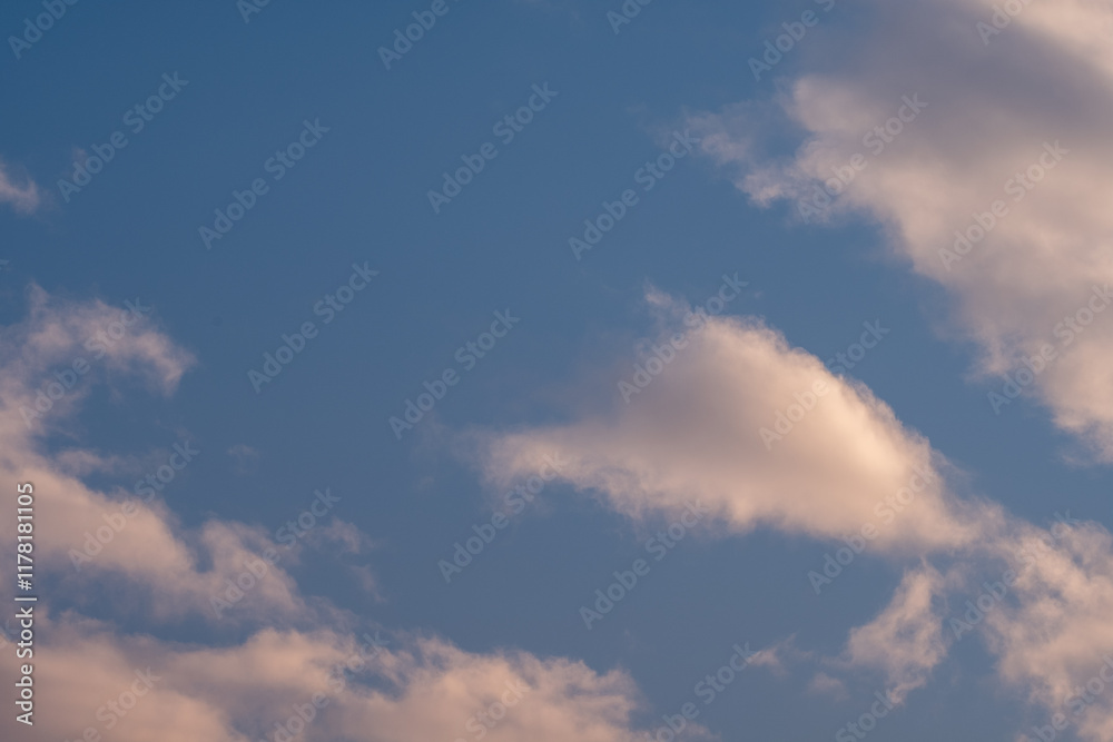Soft fluffy clouds drift across a serene blue sky bathed in the warm glow of the setting sun Soft fluffy clouds drift across a serene blue sky bathed in the warm glow of the setting sun