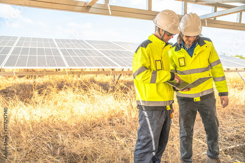 Two solar technicians in bright reflective jackets and helmets are inspecting the wiring under solar panels. One holds laptop for data logging, while the other examines electrical connections closely. photo