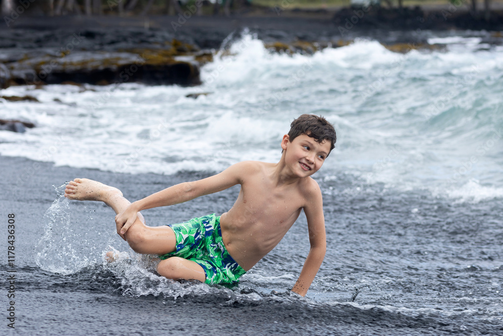 Fototapeta premium Teenager boy in colorful swimming trunks is sitting at the water's edge on the black sand, mossy rocks on the background. Black sand beach, Big Island, Hawaii, USA.