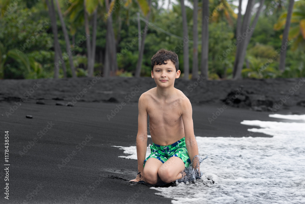 Teenager boy in colorful swimming trunks is sitting at the water's edge on the black sand, palm trees on the background. Black sand beach, Big Island, Hawaii, USA. Teenager boy in colorful swimming trunks is sitting at the water's edge on the black sand, palm trees on the background. Black sand beach, Big Island, Hawaii, USA.