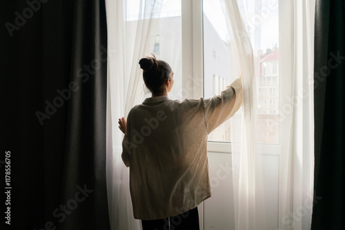 Wallpaper Mural A woman stands by a window, pulling back curtains to let in natural light and brighten her living space Torontodigital.ca