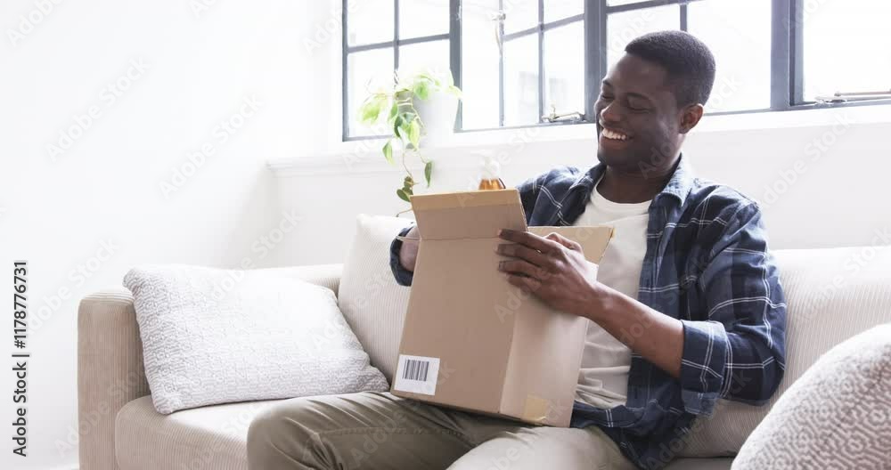 Smiling man opening gift box on couch, enjoying holiday surprise at home Smiling man opening gift box on couch, enjoying holiday surprise at home