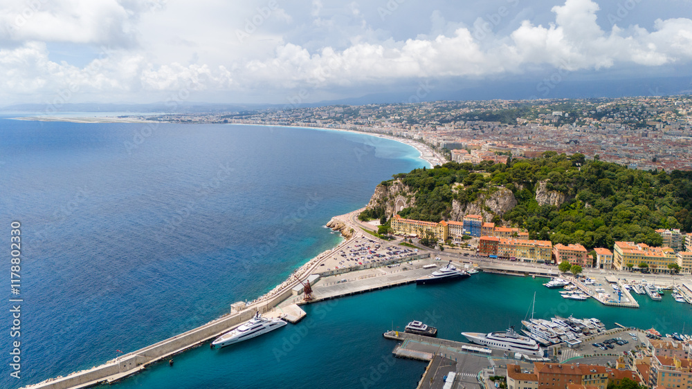Aerial view of Nice, France, showcasing the coastline with turquoise waters, luxury yachts in the harbor, and a cityscape blending with lush greenery under a partly cloudy sky Aerial view of Nice, France, showcasing the coastline with turquoise waters, luxury yachts in the harbor, and a cityscape blending with lush greenery under a partly cloudy sky