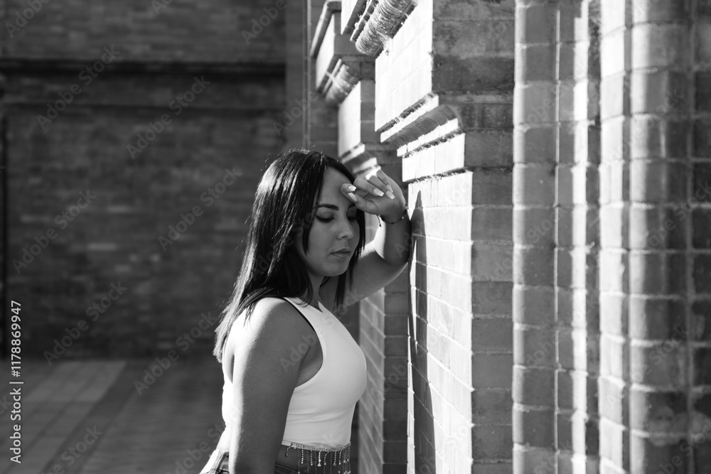 Obraz premium pretty young brunette woman rests her head on her arm on the wall and looks down. The woman is sad and depressed and feels pain and despair. She feels bad and negative. monochrome photo.