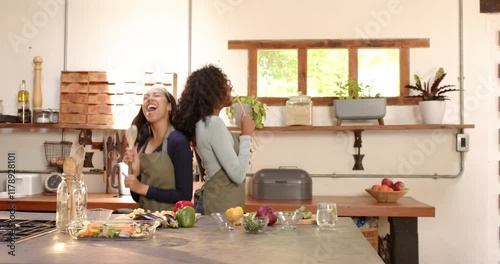 Two multiracial female friends joyfully singing and cooking together in cozy kitchen, at home