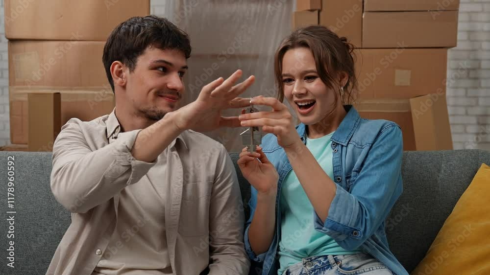Young couple moving into new apartment, space filled with cardboard boxes, man and woman sit on the sofa, man surprising woman with keys from new apartment, happy expression. Young couple moving into new apartment, space filled with cardboard boxes, man and woman sit on the sofa, man surprising woman with keys from new apartment, happy expression.