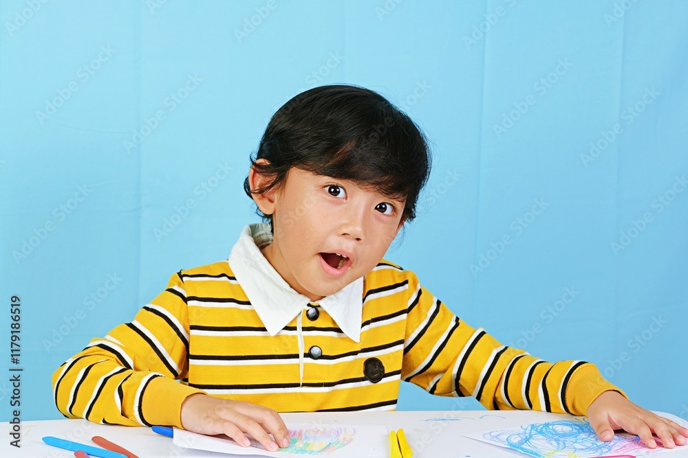 Fototapeta premium A young boy in a striped yellow shirt engaged in creative art activities with colorful crayons on a white table. Perfect for educational, childhood development, or creative learning themes.