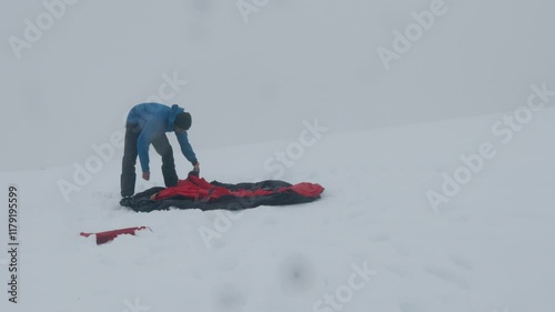 A climber in a blue outfit is setting up a bright red tarp on a snowy terrain. The visibility is limited due to snow, indicating harsh weather conditions.