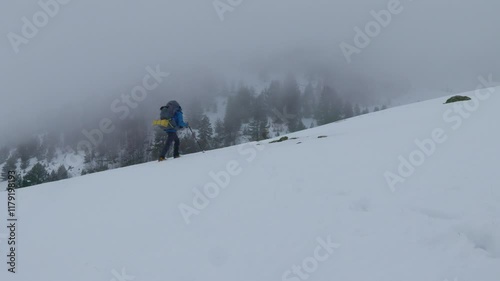 A hiker navigates through deep snow on a mountain slope amidst dense fog. The landscape is quiet, with snow covering everything in sight and visibility limited.