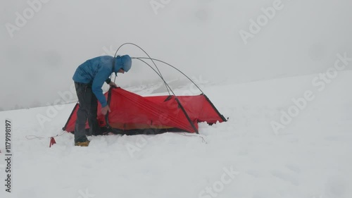 A person is assembling a red tent on a snowy landscape. Snowflakes are falling, and the atmosphere is cold, typical of a winter camping adventure in a remote area.