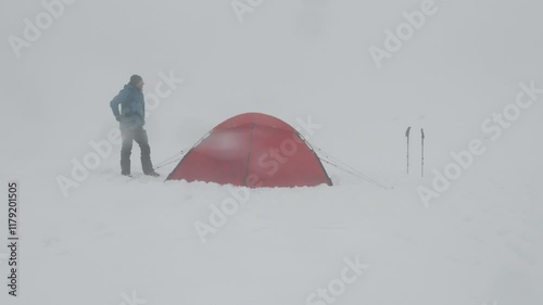 A camper is seen standing next to a bright red tent in a heavy snowstorm. The landscape is entirely covered in thick snow, creating a serene yet harsh environment.