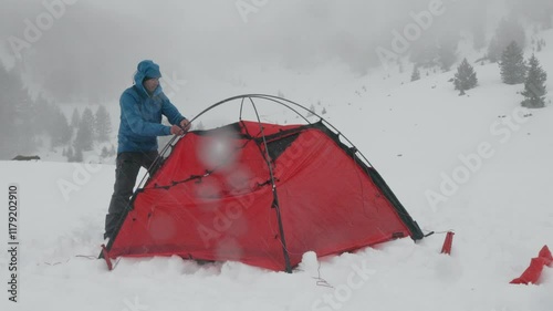 A person sets up a bright red tent in a snowy landscape. Snow falls heavily, covering the ground and creating a winter wonderland. Visibility is reduced in the frosty conditions.