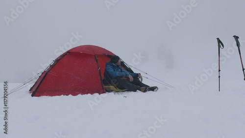 A bright red tent is set up in a deep blanket of snow, with a person relaxing nearby. Snowy mountains create a serene backdrop on a cold winter day.