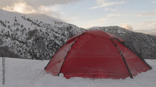 A bright red tent stands out against the snow-covered landscape, surrounded by majestic mountains under a colorful sunset sky. A person get out of tent to enjoy the view.