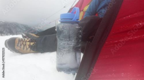 A person rests on the snowy ground in the mountains, equipped with a water bottle and wearing sturdy boots. Colorful gear contrasts against the white snow and grey sky.