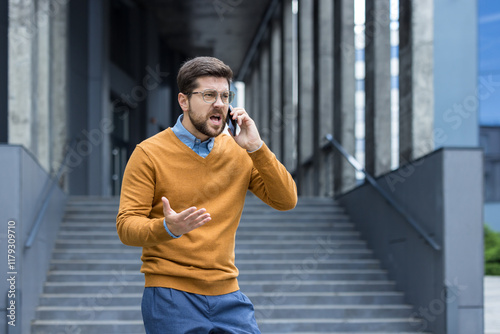 An angry young man is standing outside an office building and is talking on the phone while gesturing worriedly