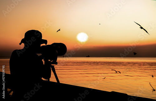 Fototapeta Naklejka Na Ścianę i Meble -  Silhouette of a photographer using  camera with large telephoto lens for shooting seagulls in the evening on sunset background. Vacation at Bangpu recreation center, Thailand the lifestyle of  hobbies