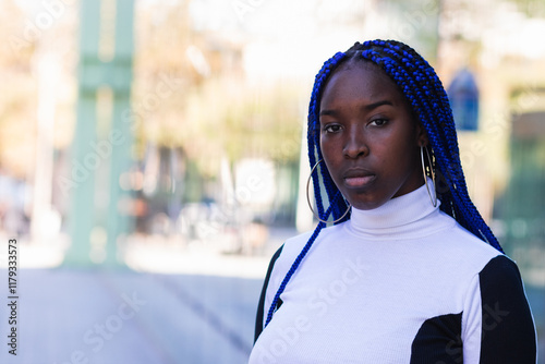 Calm African American female with blue braided hair standing in city and pensively looking at camera photo