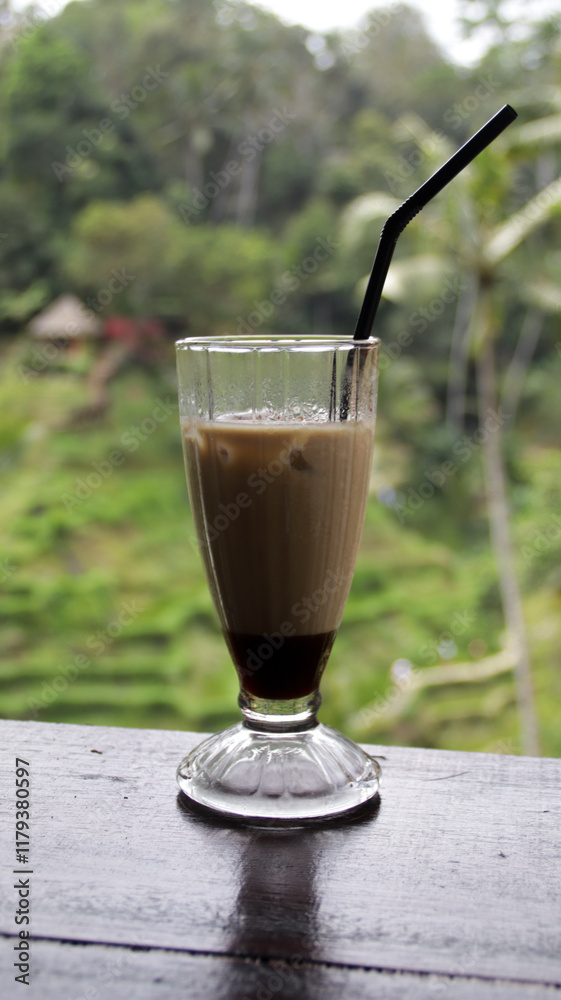 Iced latte in a tall glass with a black straw on a wooden table with blurred tropical greenery and rice terrace view in Bali Iced latte in a tall glass with a black straw on a wooden table with blurred tropical greenery and rice terrace view in Bali