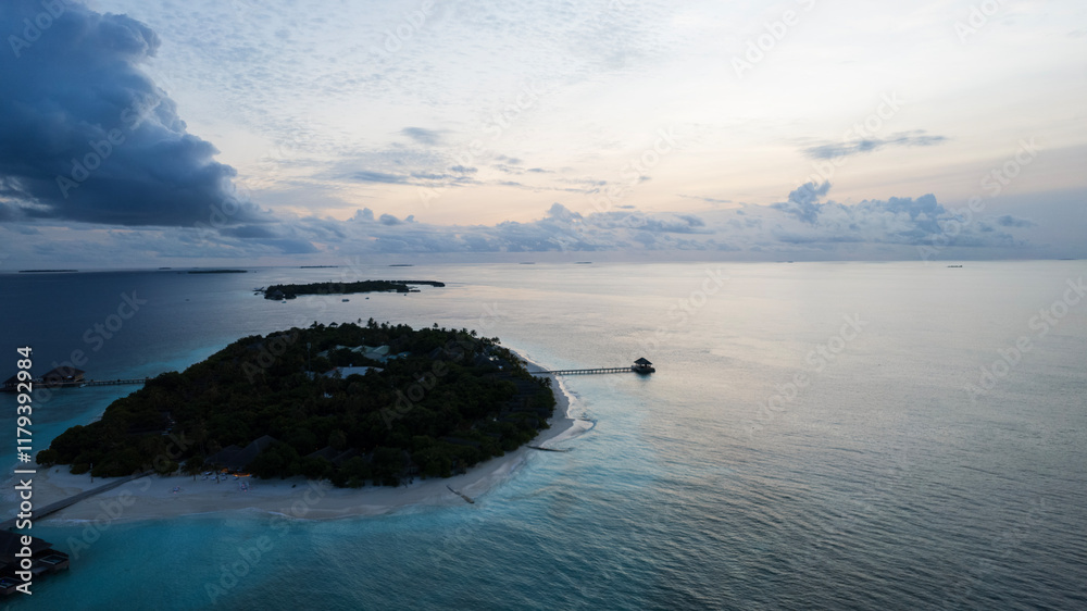 Top view of the sunset over an island in the ocean. Large dark clouds with a orange tinge. Turquoise ocean water, white sand of the beach. Green palm trees. Villas on the water. Water is like a mirror